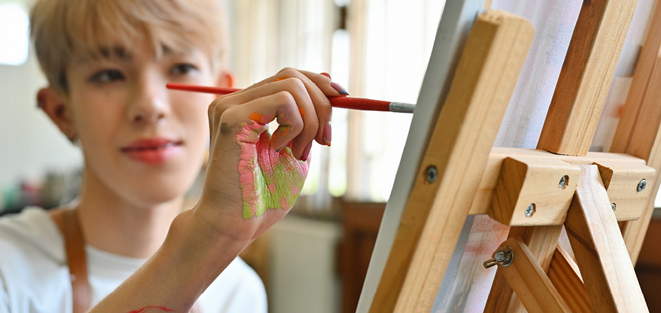 A close-up of a young artist with paint-smeared hands, focused on a canvas, applies vibrant colors with a brush. The expression reflects deep concentration and passion for their craft