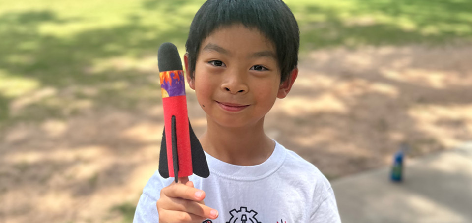 Young male student wearing a white shirt is holding a red rocket with black fins